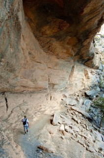 Local hiker walking under a rock overhang on the path to the abandoned settlement of Sap Bani Khamis on the western flank of the Grand Canyon of Oman in the Wadi Nakhar at the foot of the Jebel Shams ...
