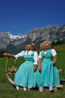 Two straw dolls or figures in dirndl dresses sitting on a sledge with long horn-shaped runners on a meadow