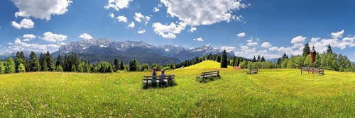 Hikers relaxing on a bench in the middle of a flowery meadow with panoramic views towards the Wetterstein Massif with the Alps and Zugspitze Mountain