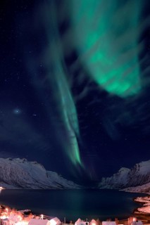 Aurora over a fjord with snow-covered mountains and an illuminated village