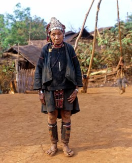 Elderly woman with a whistle standing in front of bamboo huts in an Akha village