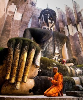 Praying monk neeling in front of the hand of the Buddha statue Phra Achana which is decorated with gold leaf