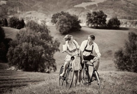 Man wearing leather pants and a woman wearing a dirndl on old bicycles within a natural landscape