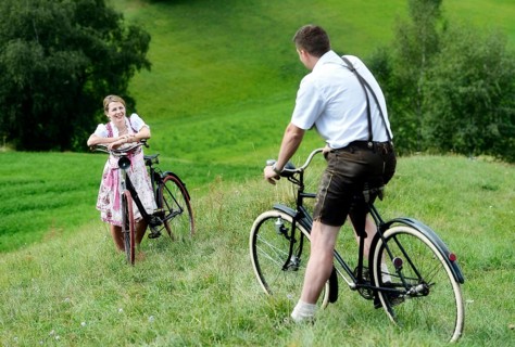 Man wearing leather pants and a woman wearing a dirndl on old bicycles within a natural landscape