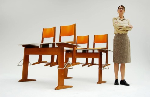 Strict female teacher standing next to old school desks with orange chairs