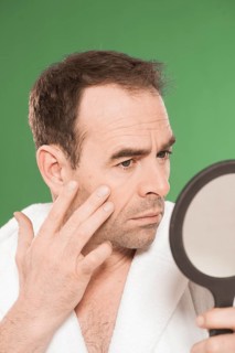 Man wearing a bathrobe observing himself critically in a hand mirror