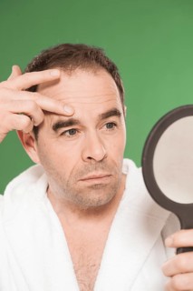 Man wearing a bathrobe observing himself critically in a hand mirror
