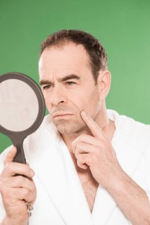 Man wearing a bathrobe observing himself critically in a hand mirror
