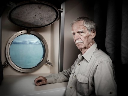 Elderly man stands in ship's cabin with a thoughtful expression
