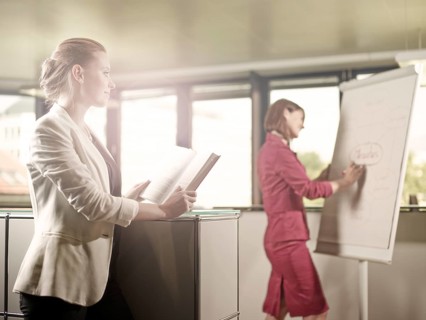 Woman reading a book and looking towards her colleague who is writing on a flip chart