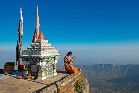 Sadhu or holy man sitting with his hands folded in prayer on the precipice in front of a Hindu temple shrine with prayer flags