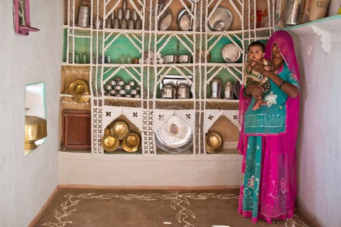 Young woman in colourful sari proudly standing with a child on her arm in front of a traditional kitchen shelf built from wire and sticks