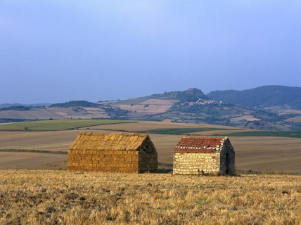 Bales of straw stacked in the shape of a house next to a little stone house