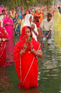 Woman wearing a red sari with offerings standing in water during the Hindu Chhath Festival