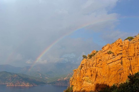 The typical bizarre red rocks of the Calanche of Piana and the mediterranean sea at the Gulf of Porto in the background below a rainbow and some clouds. The Calanche of Piana is in the western part of...
