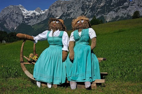 Two straw dolls or figures in dirndl dresses sitting on a sledge with long horn-shaped runners on a meadow