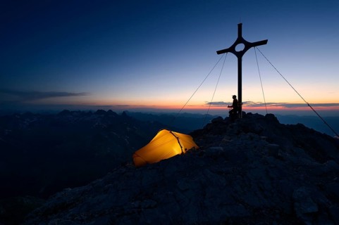 Mountaineer with a tent at the summit cross on Großer Krottenkopf Mountain at dusk