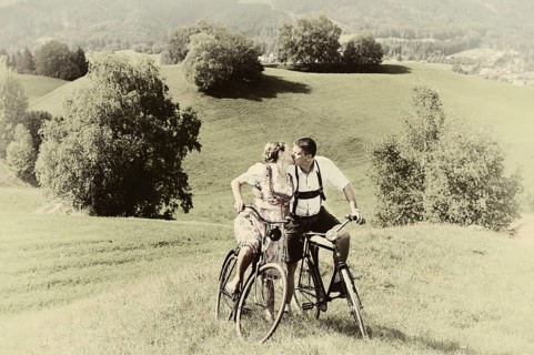 Man wearing leather pants and a woman wearing a dirndl kissing on old bicycles within a natural landscape