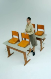 Strict female teacher standing at old school desks with orange chairs