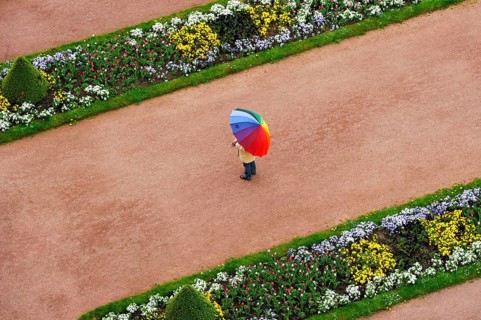 Person with umbrella in rainbow colors on a red gravel path between flower beds in the castle gardens