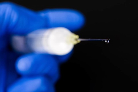 A hand with a blue medical glove is holding a syringe with a hypodermic needle and a drop of medicine