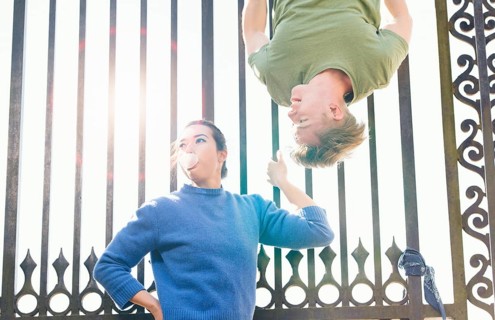 Young man trying to impress indifferent girl by hanging upside down on an iron gate