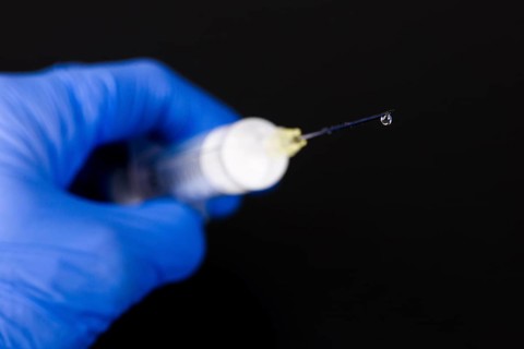 A hand with a blue medical glove is holding a syringe with a hypodermic needle and a drop of medicine