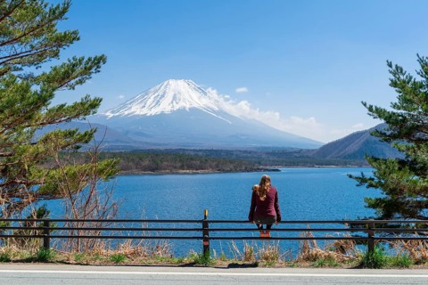 Young woman sitting on a railing next to a road and looking across the lake to the volcano Mt Fuji