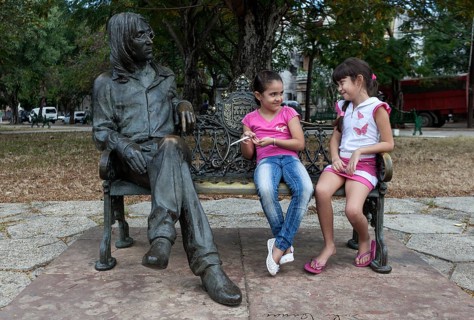 Two girls sitting on a bench next to a bronze sculpture of John Lennon