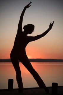 Woman with backlighting doing a yoga pose on a wooden pier at a lake in front of the setting sun