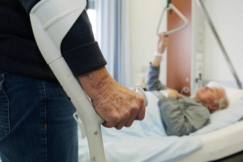 Elderly woman standing on a crutch in front of an elderly man lying in a hospital bed