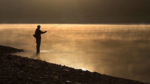 A fly fisherman standing on the shore of the Obernau reservoir at sunrise