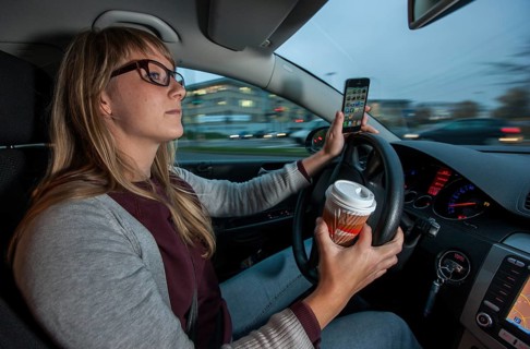 Young woman driving a car in the city centre while holding her cell phone and a cup of coffee at the same time