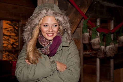 Smiling young woman wearing a green parka standing with her arms crossed in front of a hiking hut decorated for Christmas