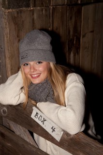 Smiling young woman wearing a hat and scarf standing at the gate of a horse's stall