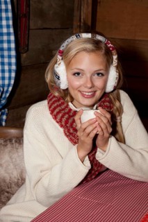 Smiling young woman wearing ear warmers and a scarf sitting at the table of a hiking hut and drinking a hot drink