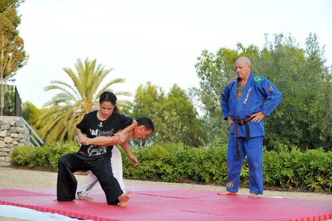 Woman and a man doing martial arts training on a sports mat in a park while being observed by their trainer