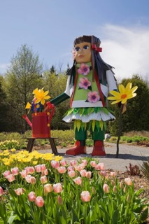 A tall native girl like doll stands next to a patch of pink and yellow tulips in the Greeting garden at the "Route des Gerbes d'Angelica" garden at springtime