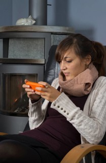 A young woman of Indonesian origin is sitting in front of a wood burner working on a smartphone