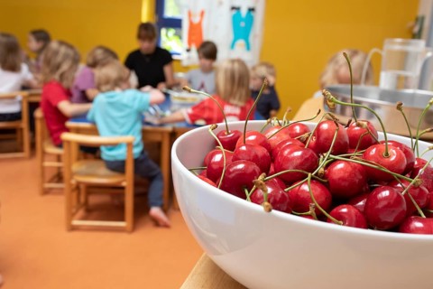 A bowl of cherries stands in the dining room in the kindergarten and children sit at the table to eat