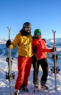 Two skiers with ski helmets and skis standing on the ski slope in front of a mountain panorama