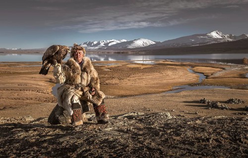 Mongolian eagle hunter posted with lake in front of landscape with lake and snow-covered mountains