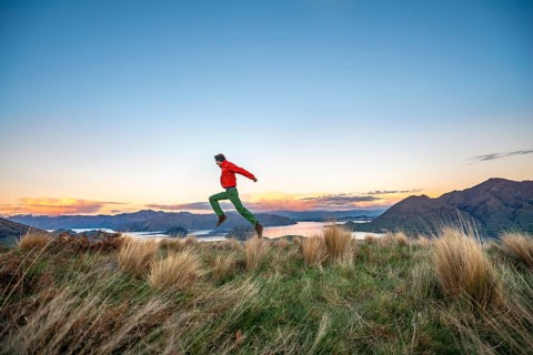 Hiker jumps into the air, panoramic view of Wanaka Lake and mountains at sunset
