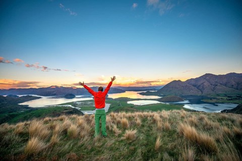 Hiker stretches his arms in the air, panoramic view of Wanaka Lake and mountains at sunset