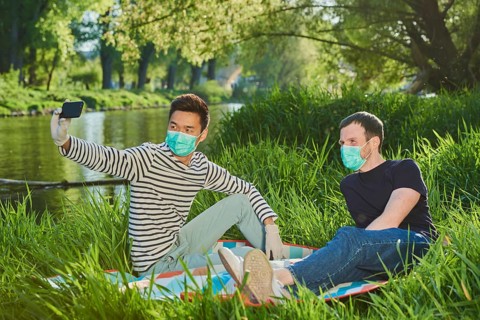 Two men with face masks sitting in a meadow by the river