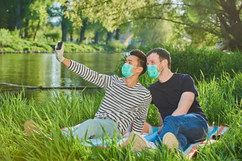 Two homosexual men with face masks sitting in a meadow by the river