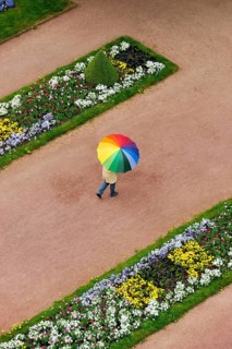 Person with an umbrella in rainbow colours on a red dirt road between flowerbeds in the Palace Gardens