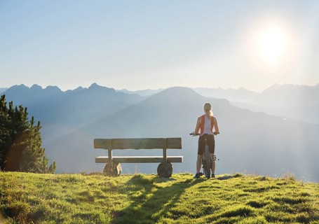 Mountain biker stands with MTB next to a wooden bench and looks into the wide light-flooded mountain landscape