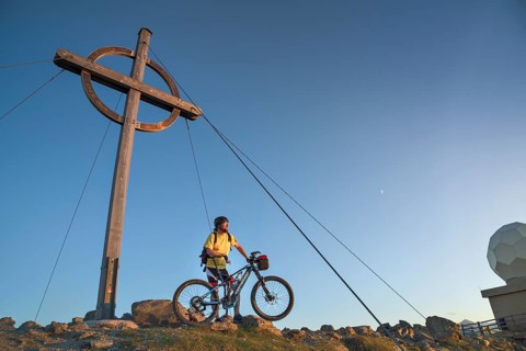 Mountain biker stands with e-MTB next to the summit cross of the Patscherkofel in the warm evening light