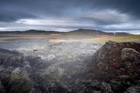 Black solidified lava in green plain with steam